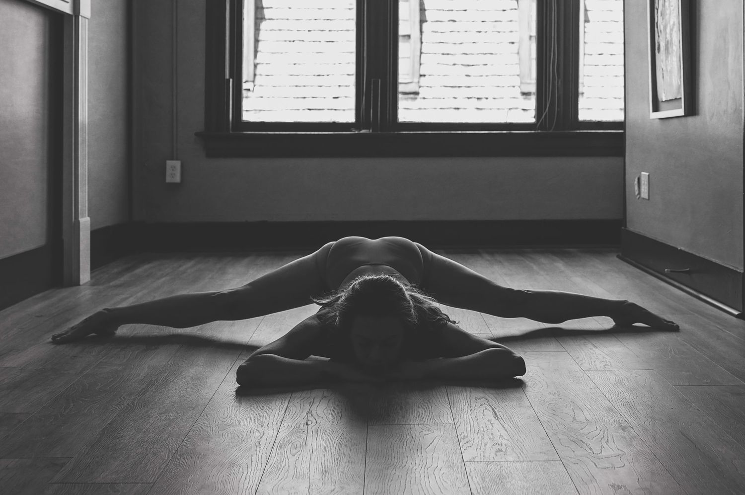 A person lies stretched out in a yoga pose on a hardwood floor in front of a bright window.