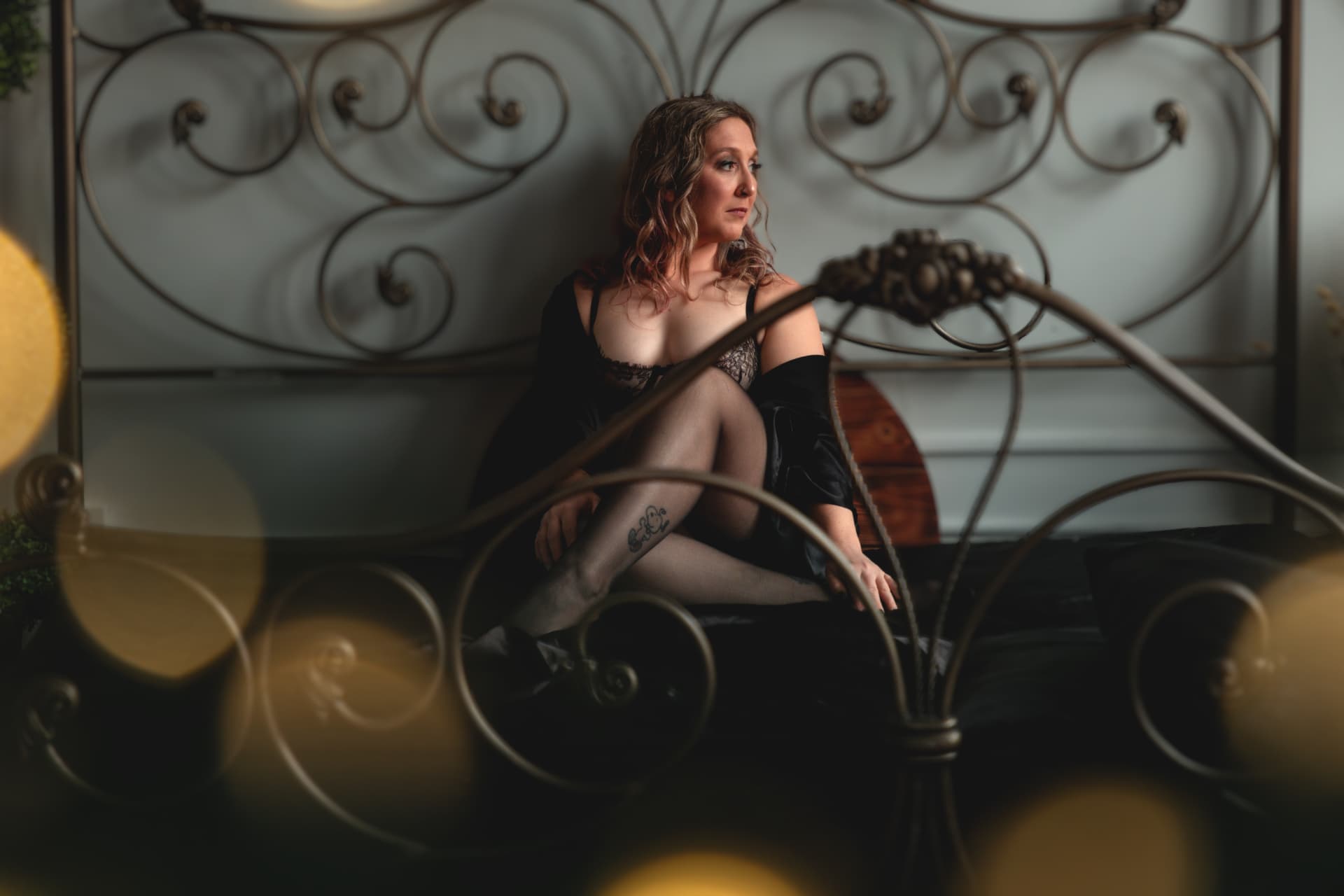 A woman in black lingerie and a robe sits on an iron bed in warm, moody studio light.